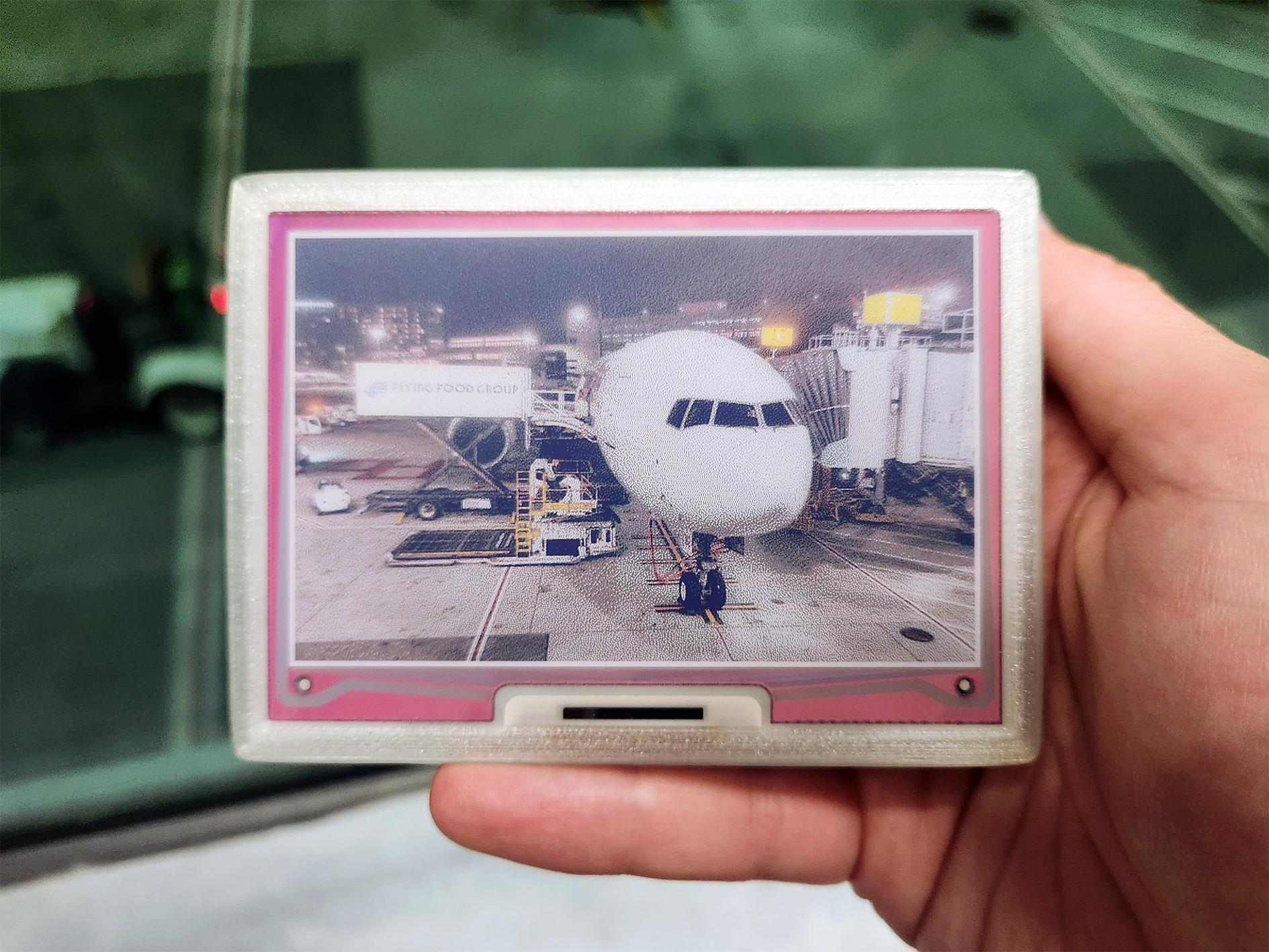 A photo of a hand holding eink.cam. On the camera is the same photo from earlier, showing the plane being loaded.